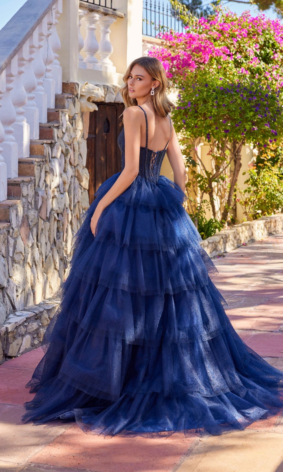 A woman stands outdoors on a stone path near flowers, wearing the Long Prom Dress: Juliet JT2611N, a dark blue tiered glitter tulle ball gown with a beaded corset bodice.